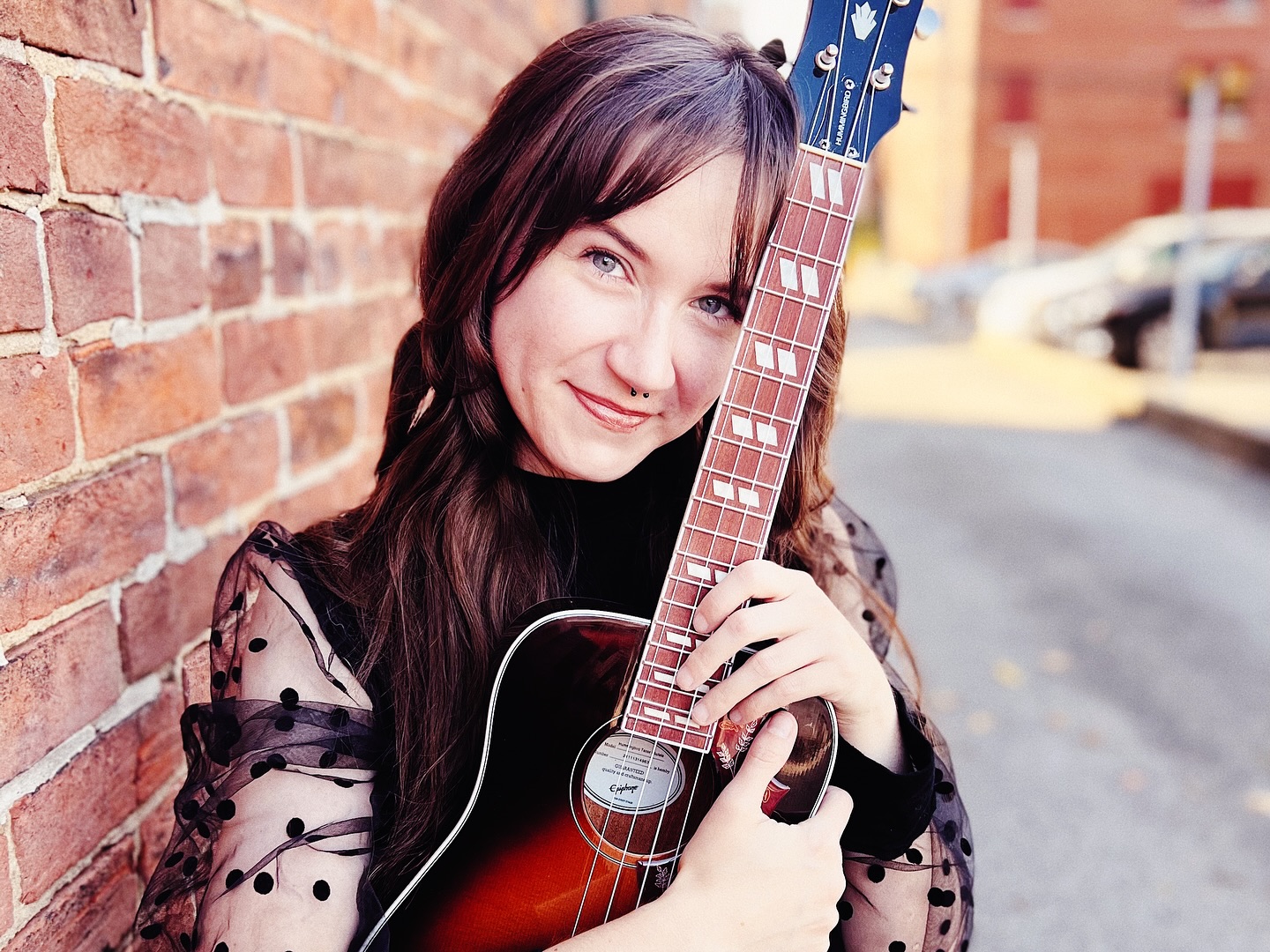 Close-up portrait of a smiling musician holding an acoustic guitar against a brick wall.
