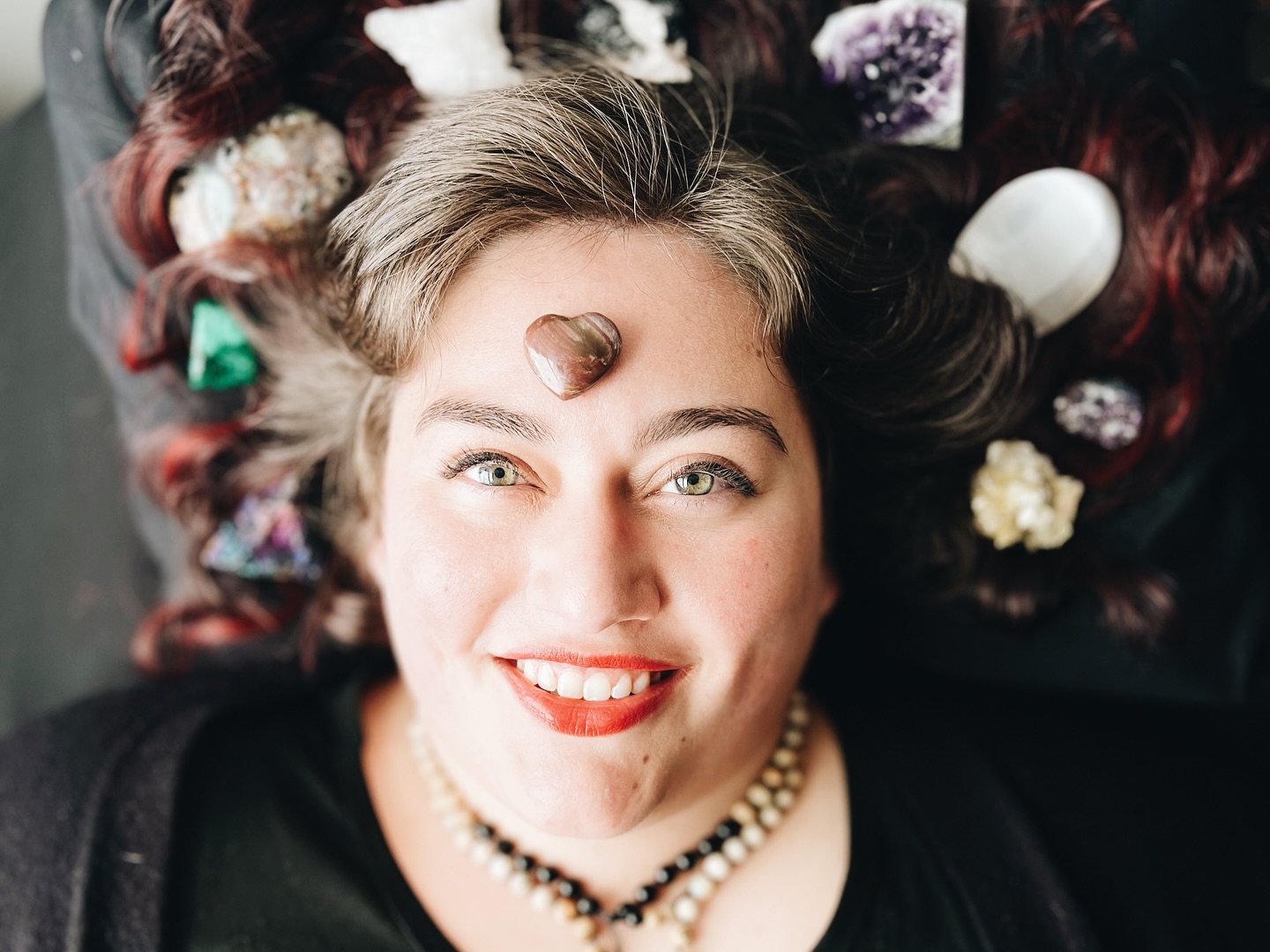 Overhead close-up of a smiling woman with a heart-shaped stone on her forehead and crystals in her hair.