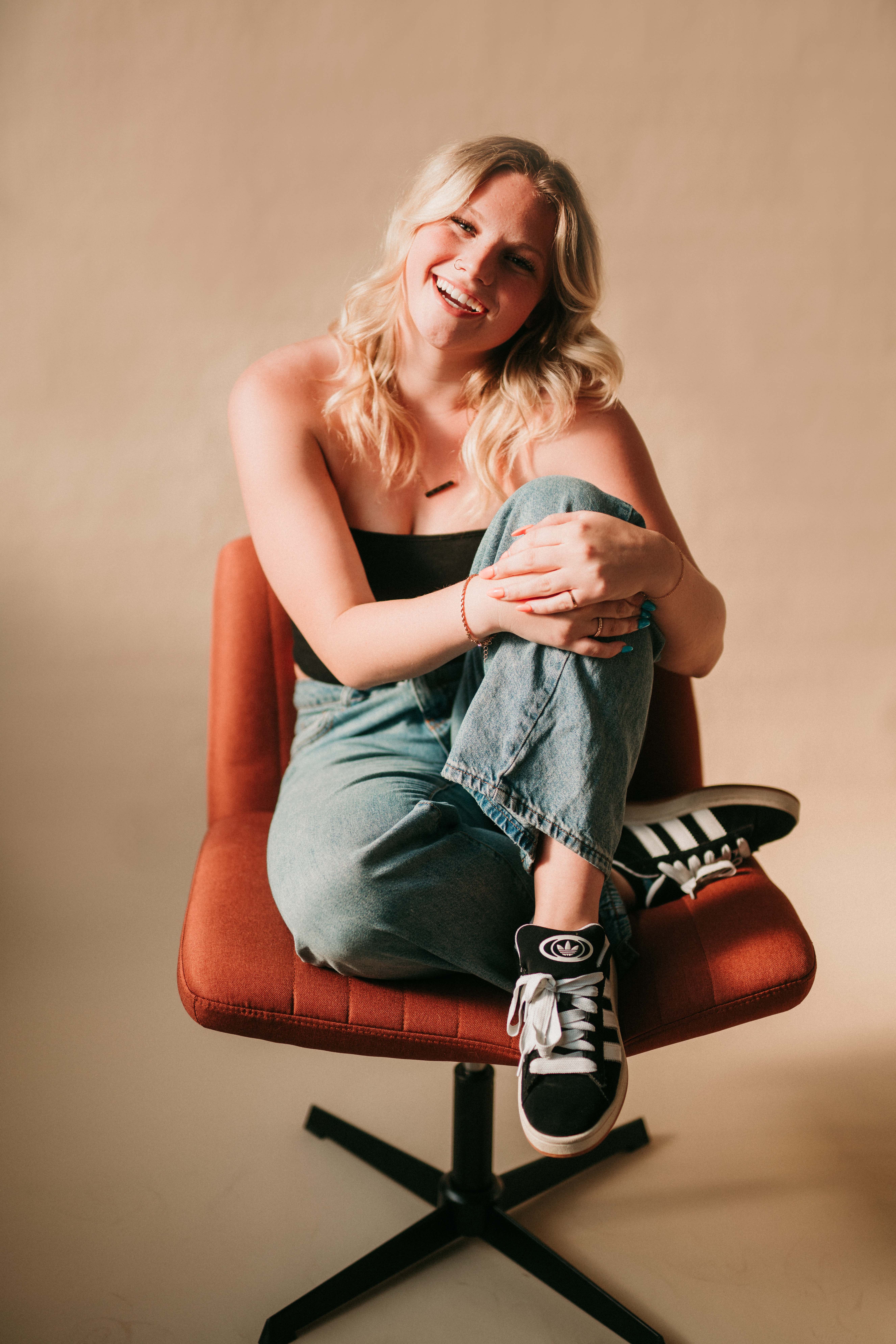 Smiling blonde senior sitting casually on an orange chair in a studio.