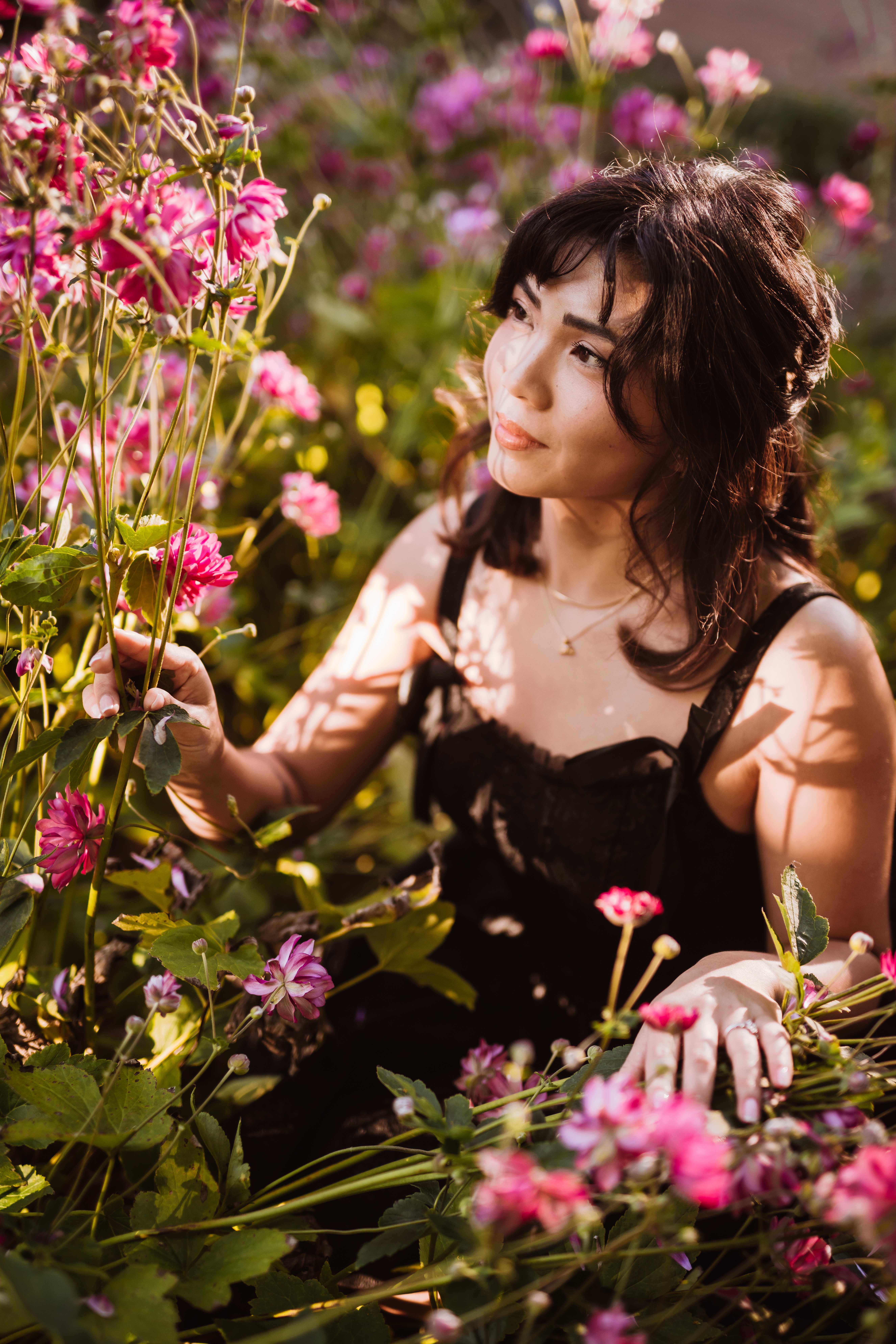 Senior with dark hair in a black dress sitting thoughtfully among pink flowers in a garden.