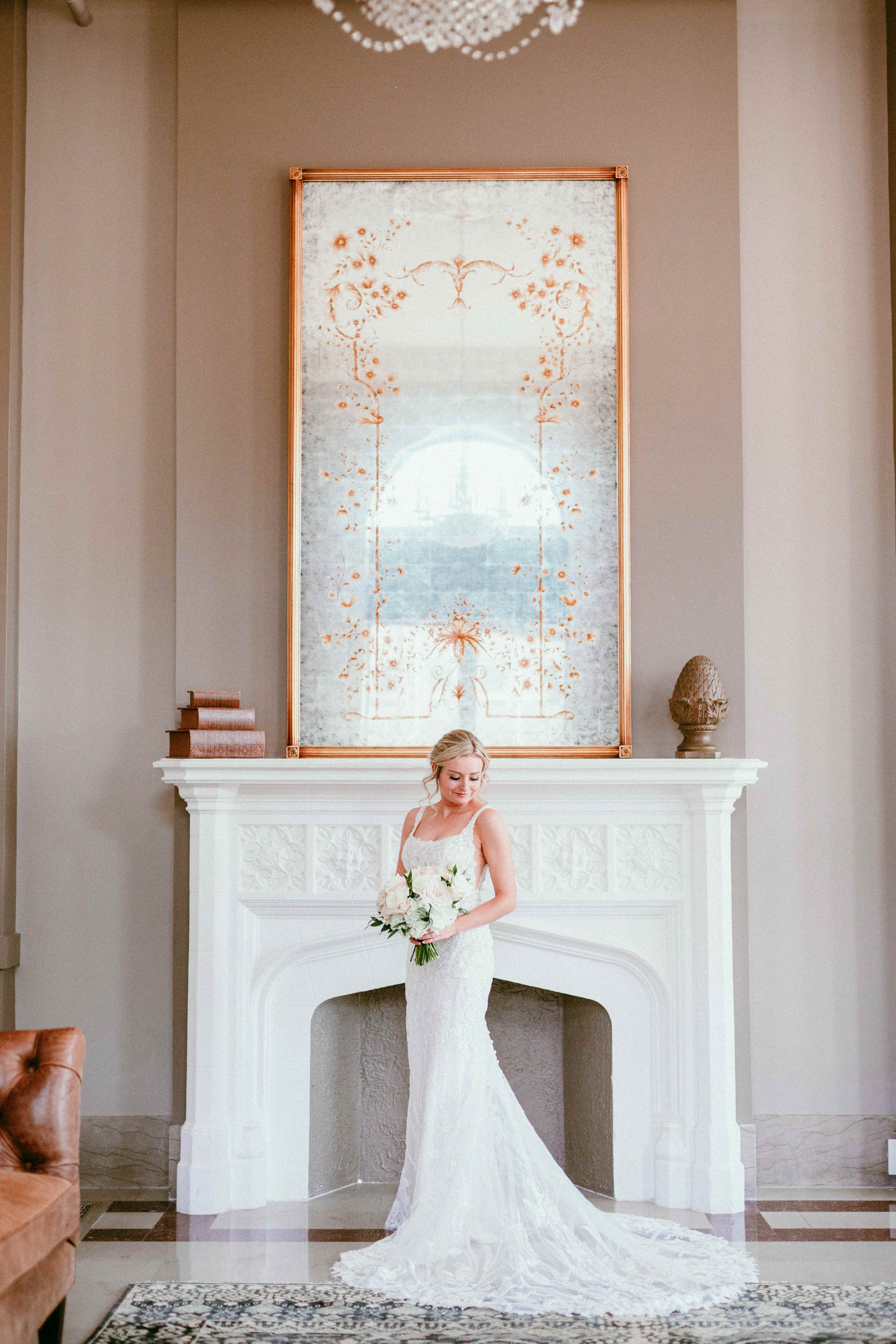 Elegant blonde bride in a lace gown posing by an ornate white fireplace.