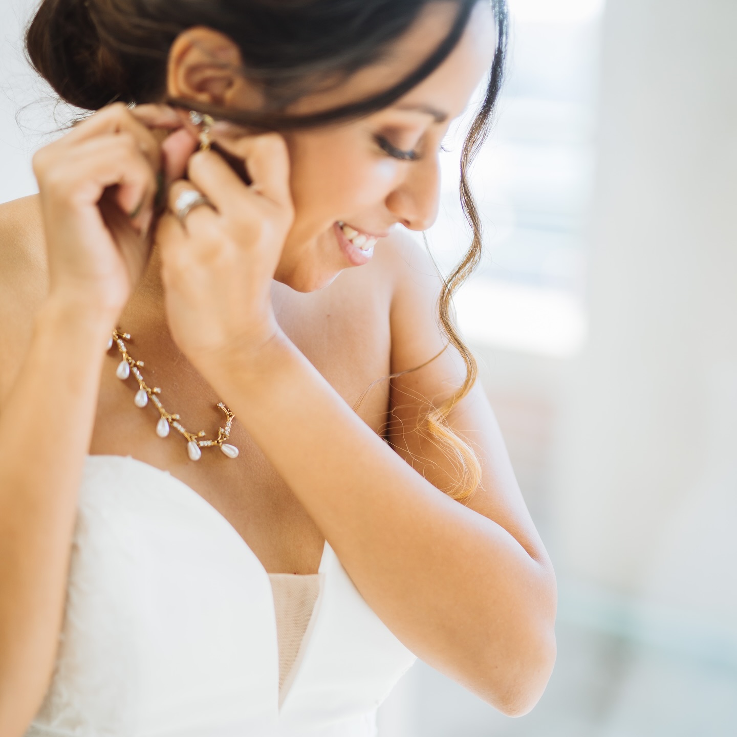 Close-up of a smiling bride putting on her earring, wearing a pearl necklace and strapless gown.
