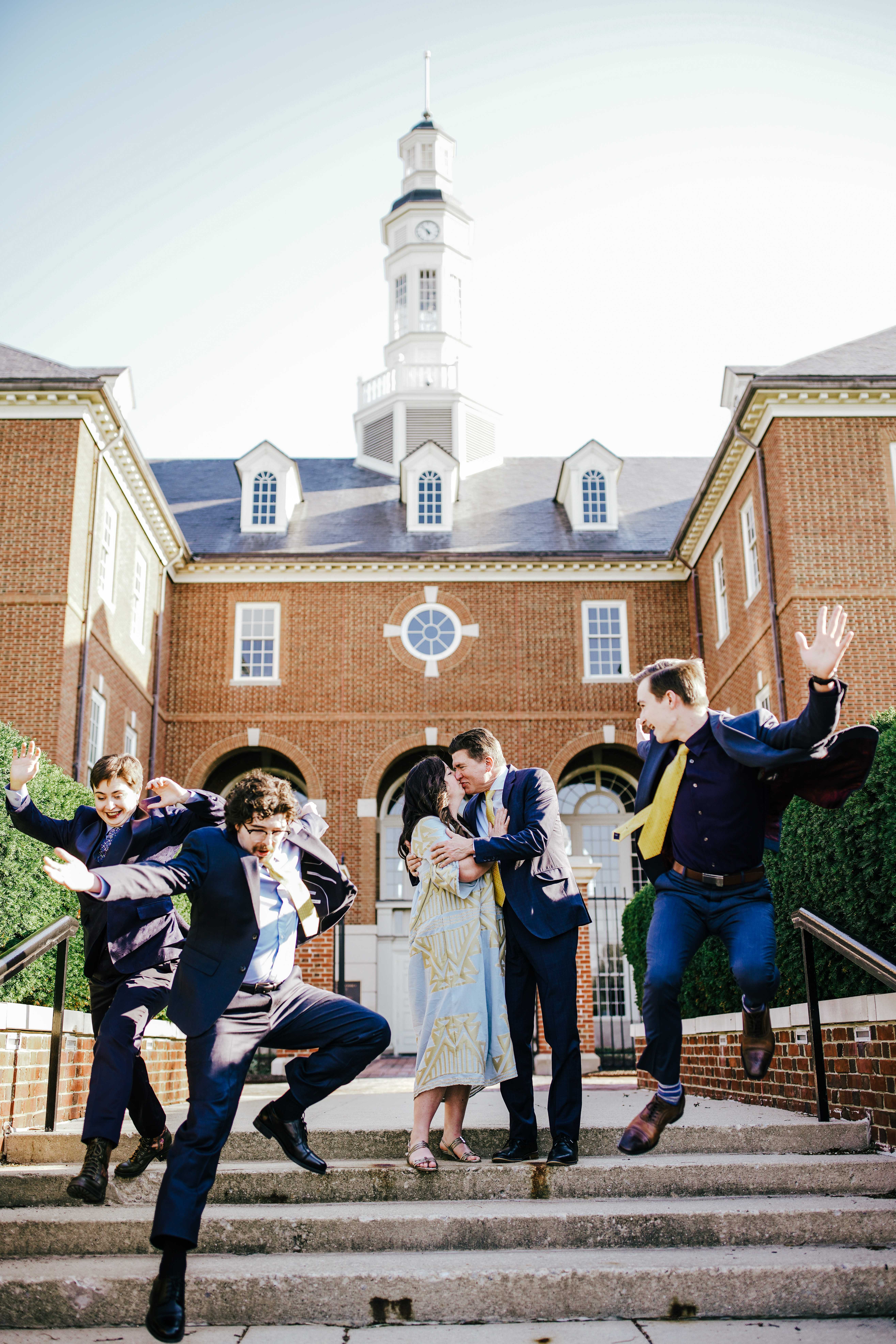 Fun wedding photo of couple kissing while groomsmen jump around them on steps.